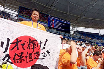 [写真]久保恒造選手へのメッセージの入った国旗を持つ荒井監督